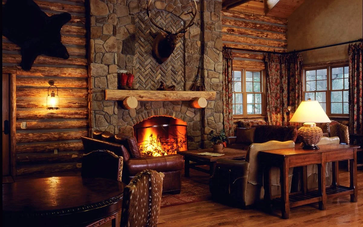The lodge great room: log walls, a river-rock fireplace with the fire lit, leather chairs and a lamp under the beamed ceiling